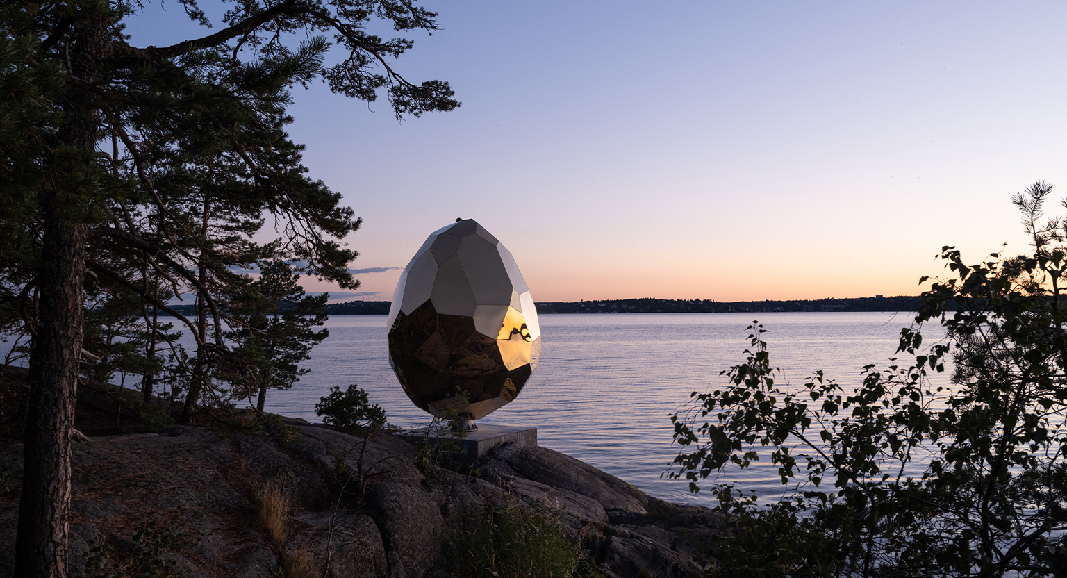 Bilden föreställer skulpturen Solar Egg i skymning. Skulpturen i form av ett gyllene ägg står på en klippa vid vattnet, solen har precis gått ner i horisonten.