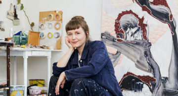 Konstnären Disa Isæus-Daggfeldt sittandes framför sitt konstverk i målarkläder