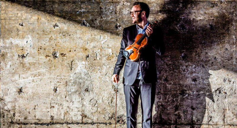 Violinisten Daniel Migdal står framför en vägg av betong och ler med sin fiol i handen
