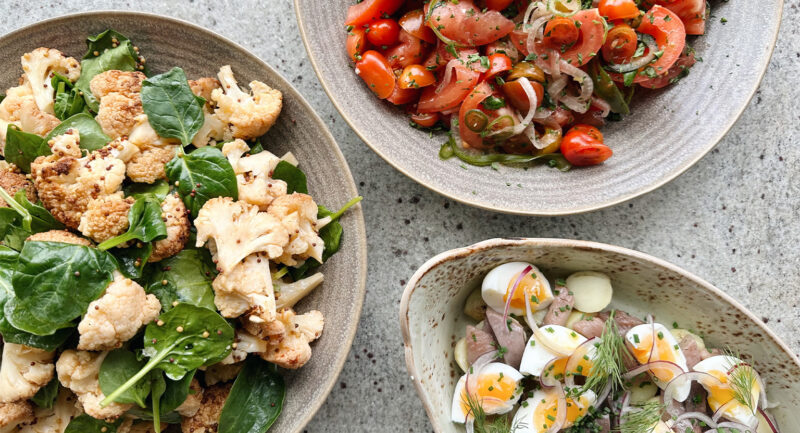 Bild på tre rätter ovanifrån. Den ena rätten föreställer en sallad innehållandes rostad blomkål. Den andra rätten är en sallad med tomater och tunt skivad lök. Den tredje rätten är en lerpottasill med matjesill, ägg, potatis och dill.