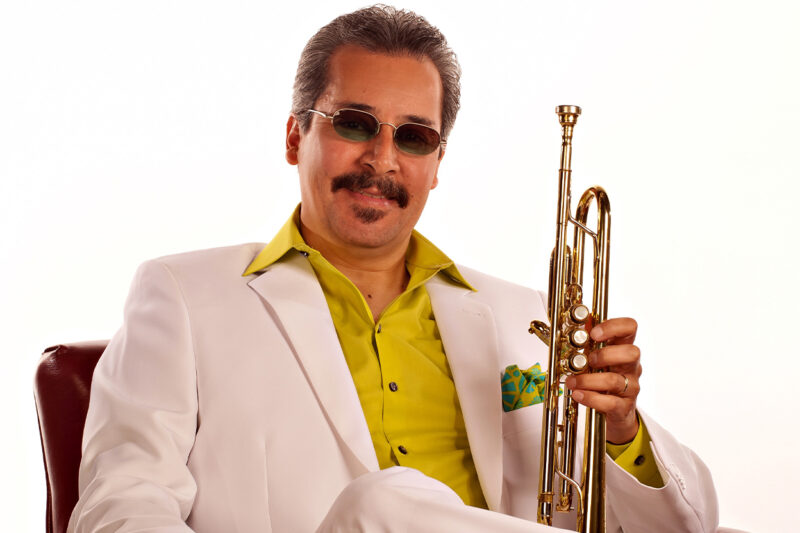 En man i vit kostym och gul skjorta sitter i en läderfåtölj och håller i en trumpet.