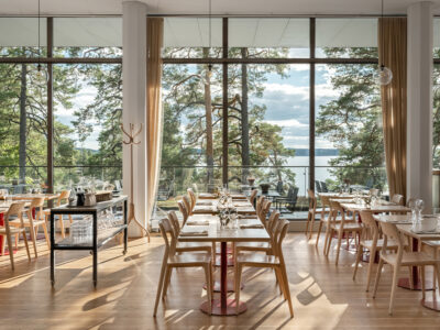 Bilden förställer Artipelag Restaurang i solljus. På bilden syns dukade bord på rad framför ett panoramafönster med utsikt över vattnet och talltopparna.