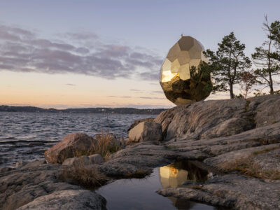 Bilden föreställer skulpturen Solar Egg i skymning. Skulpturen i form av ett gyllene ägg står på en klippa vid vattnet, solen har precis gått ner i horisonten.