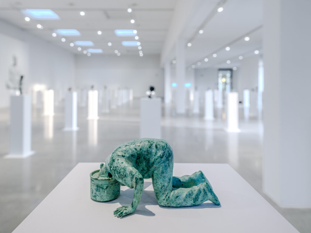 Utställningsvy med en skulptur i förgrunden. I bakgrunden finns en mängd piedestaler med skulpturer.