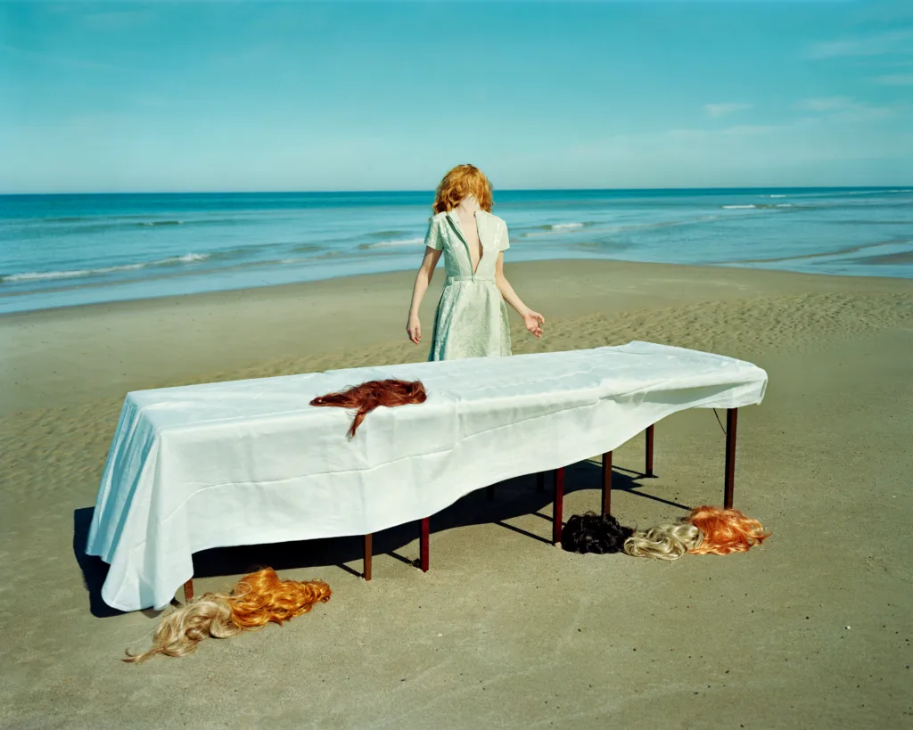 Ver av Denise Grünstein, med en kvinnokropp stående på en strand med ett bord framför henne. På bordet och på stranden ligger peruker. Kvinnan verkar ansikts- eller huvudlös, med en peruk.